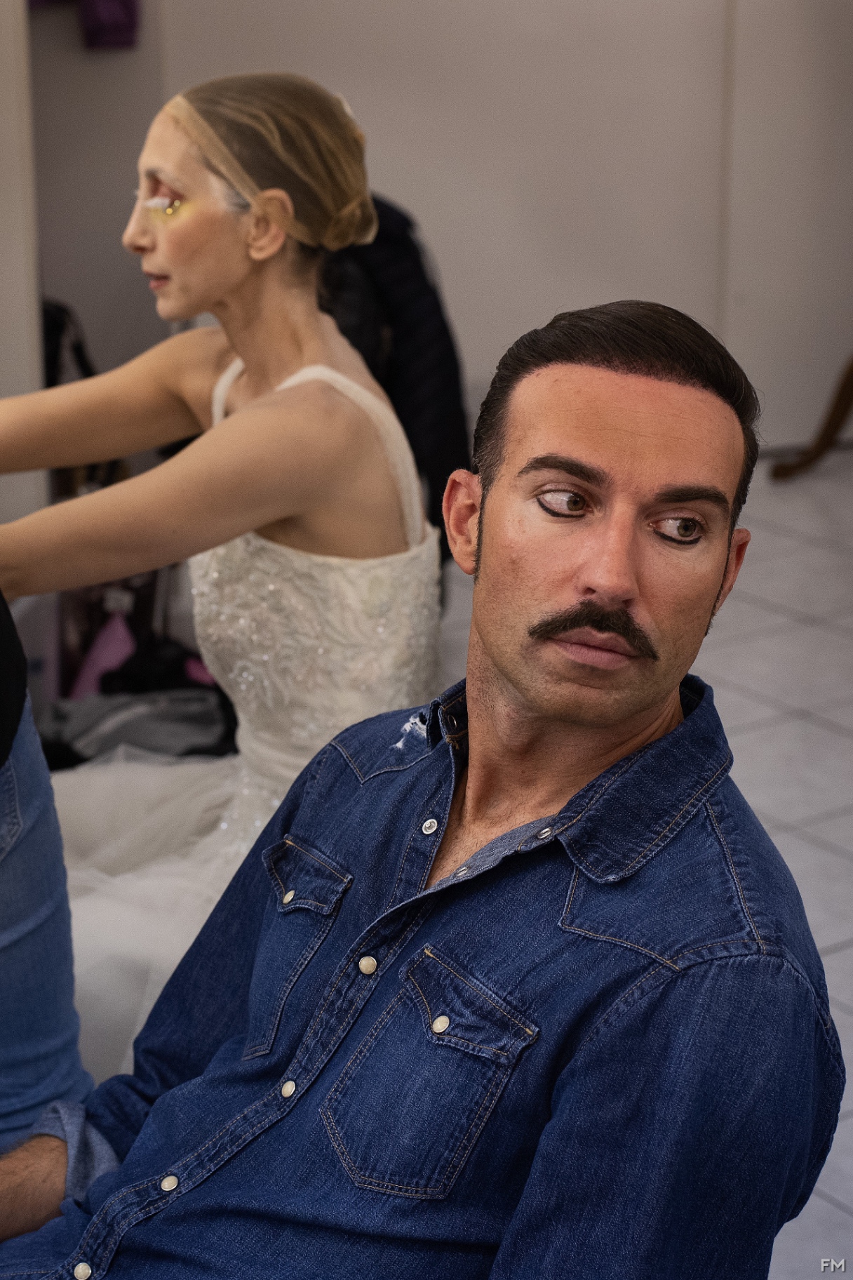 Attore con baffi disegnati e camicia di jeans in primo piano, ballerina in abito bianco sullo sfondo