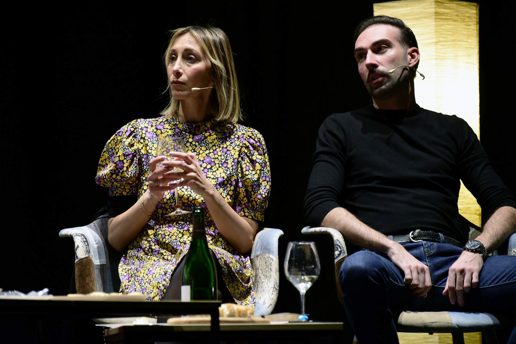 Coppia seduta fianco a fianco, lei con calice, lui in nero, sguardi opposti