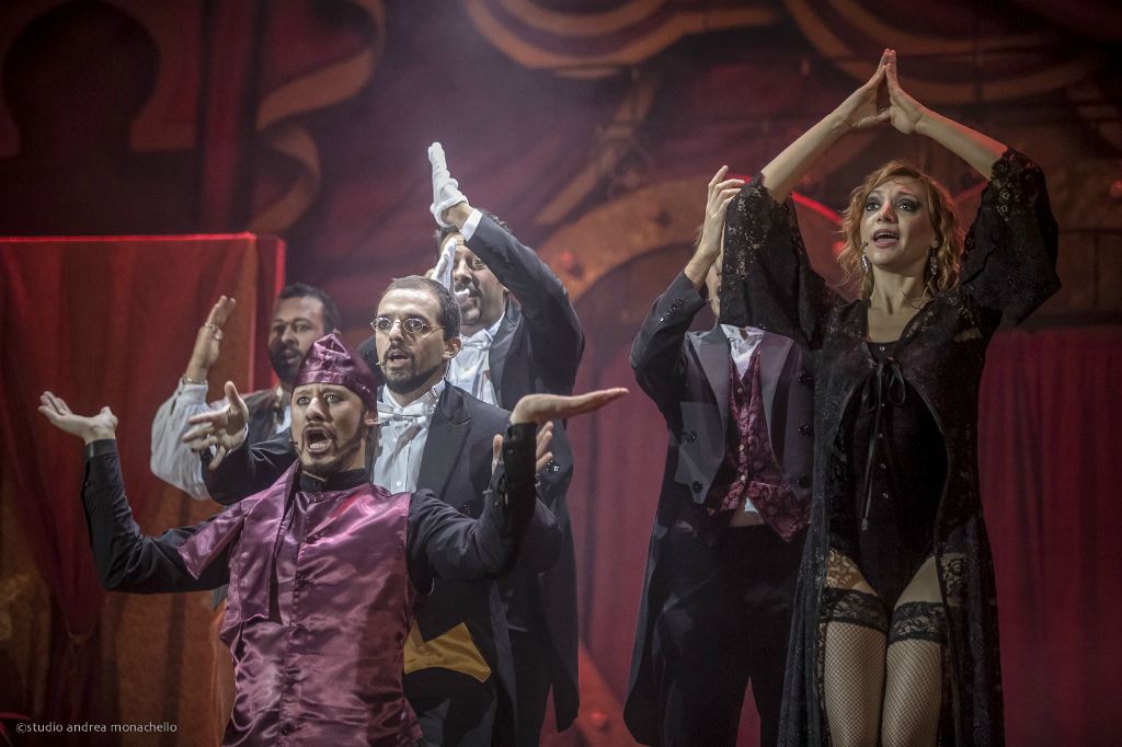 Ensemble in scena: uomo in maglia viola con bocca spalancata e donna in pizzo nero a braccia alzate