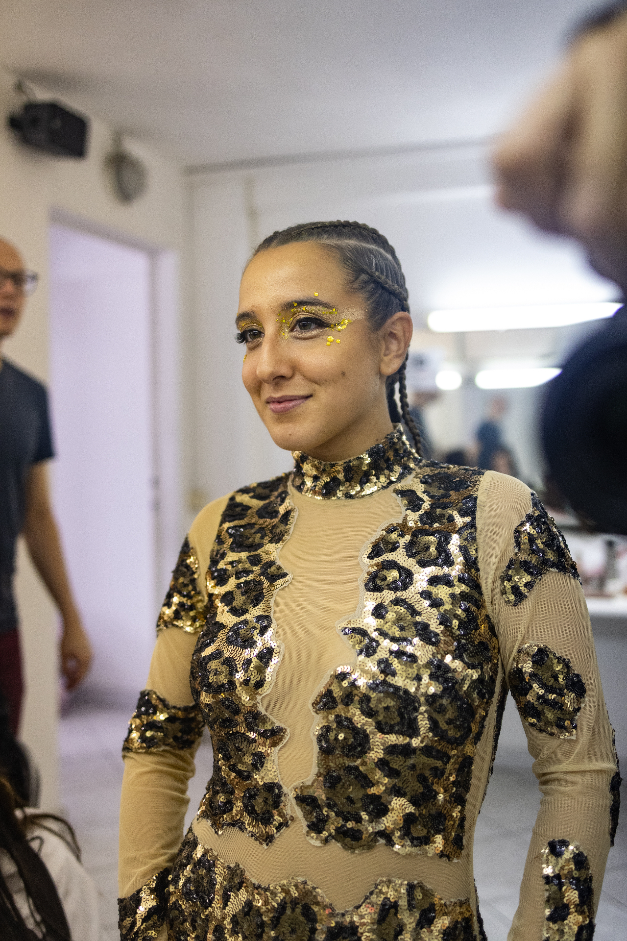 Ballerina in body leopardato dorato con trecce raccolte e glitter giallo sugli occhi sorride in camerino
