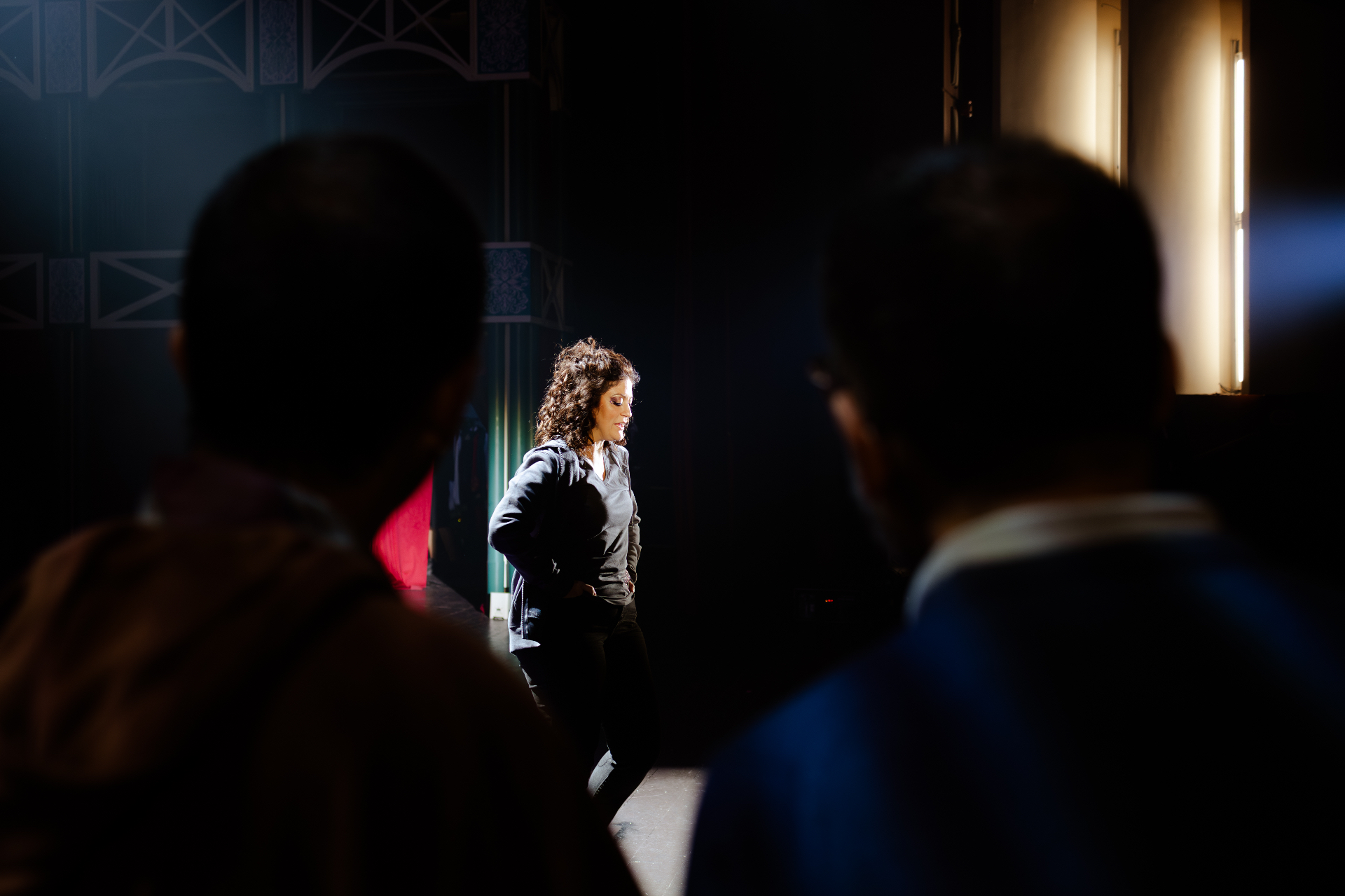 Attrice sul palco illuminato da un taglio di luce, osservata di spalle da due colleghi in ombra