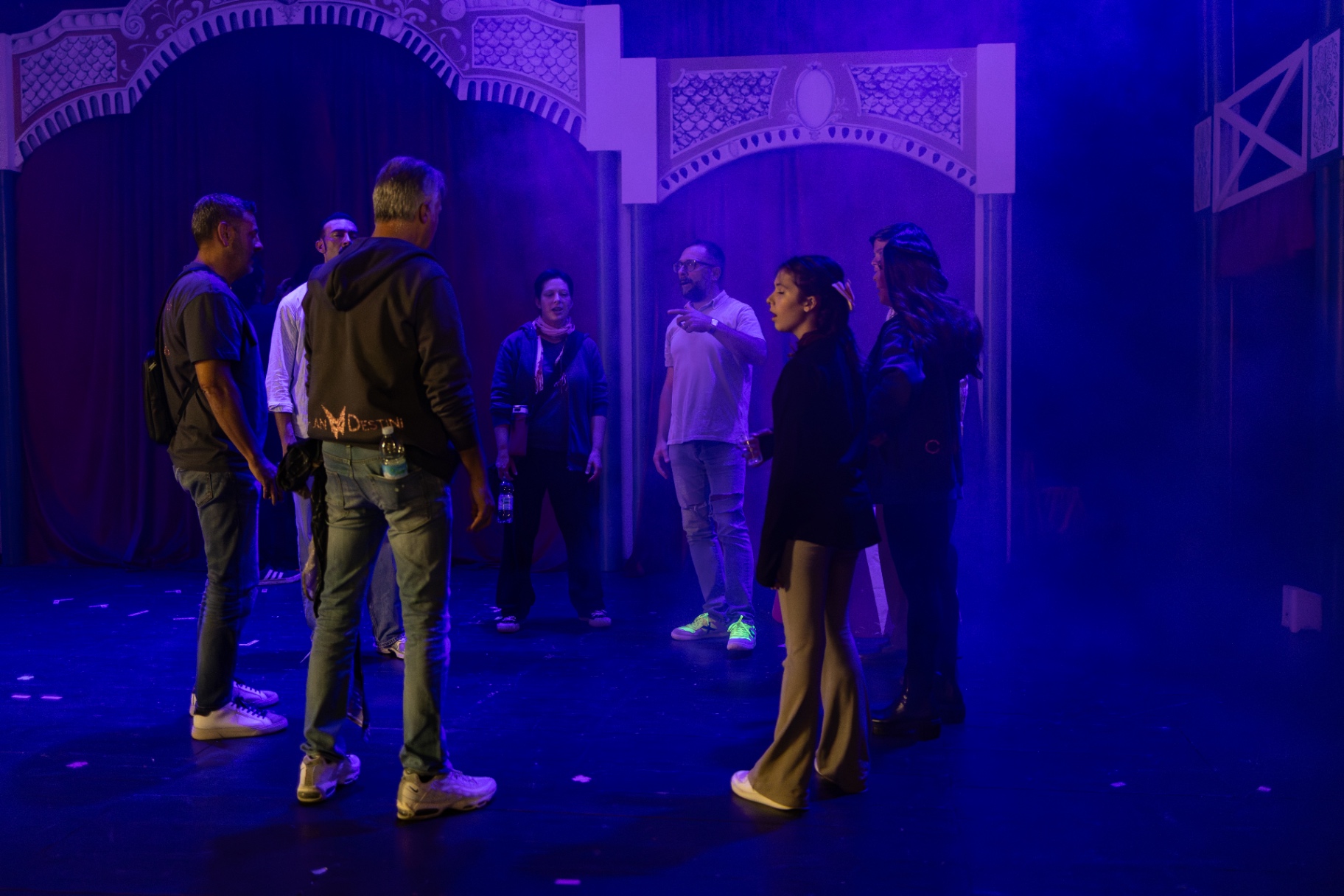 Cast riunito sul palco sotto luci blu davanti al sipario rosso per un ultimo confronto prima dello spettacolo