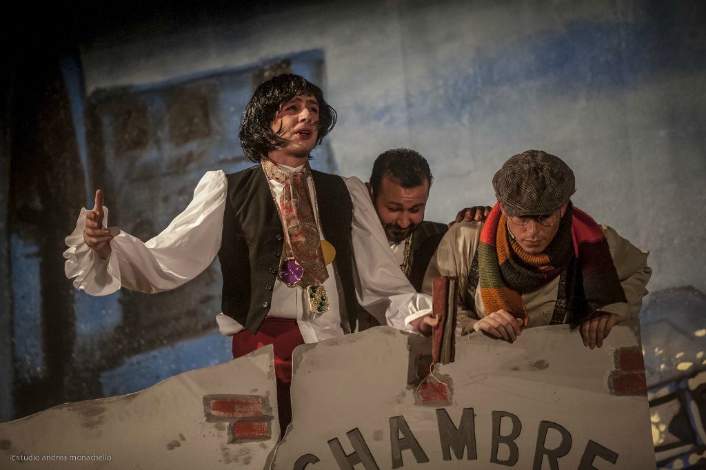 Tre attori in costume Ottocento parigino accanto a un muro dipinto blu con scritta Chambre