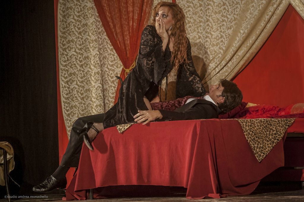 Donna in pizzo nero su un letto rosso con uomo riverso sulle sue ginocchia