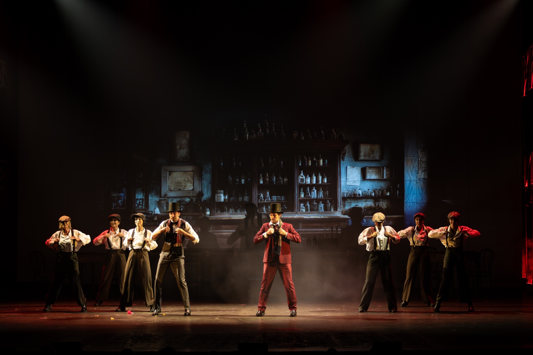 Cast in eleganti bretelle e cappelli neri in coreografia sincrona davanti a un bar dipinto