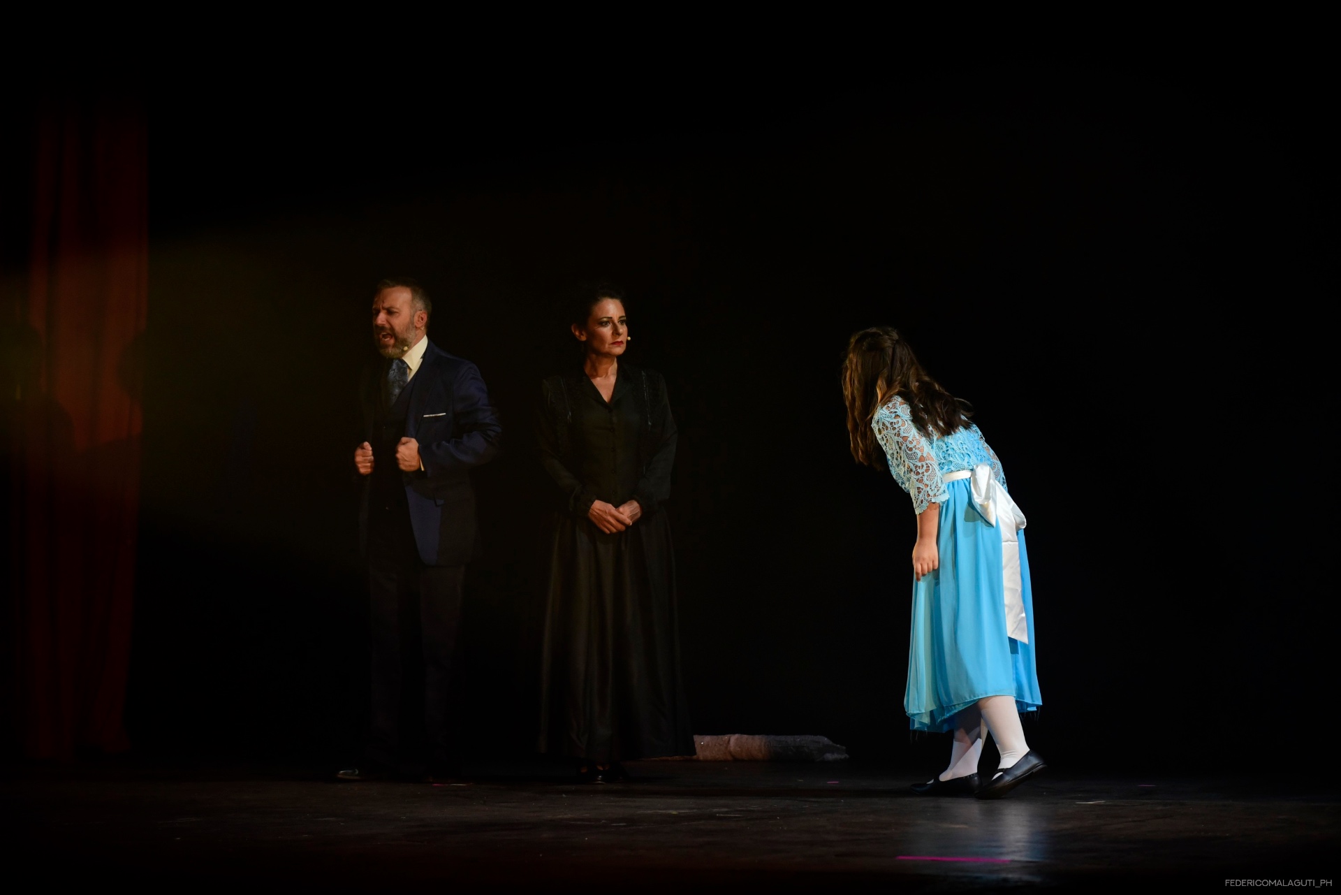 Uomo, donna e giovane in scena con luci basse e sipario rosso sullo sfondo