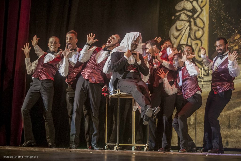 Coro maschile in gilet rosso con braccia alzate e mani aperte attorno a un cantante seduto