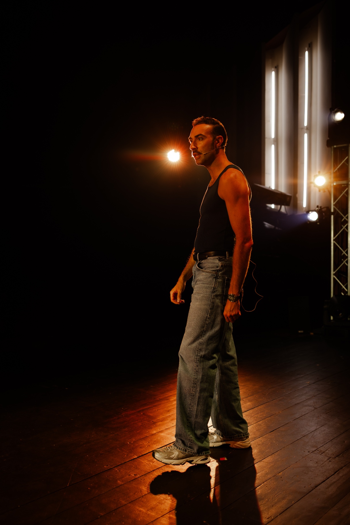 Attore in canottiera nera e jeans in piedi su un palco buio illuminato da tagli di luce arancione e neon bianchi