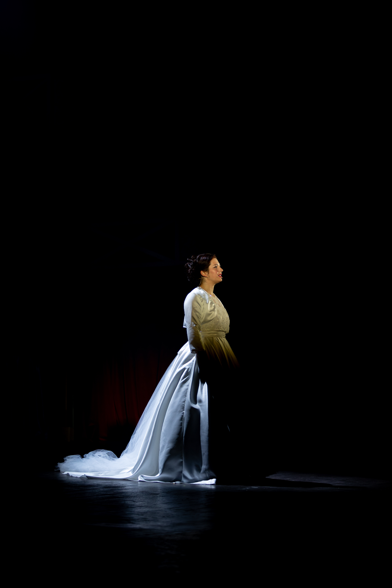 Donna in abito da sposa bianco satinato in piedi sotto un fascio di luce sul palco buio