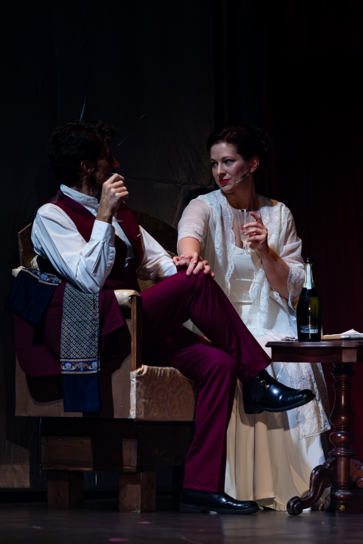 Uomo in gilet bordeaux e donna in vestaglia bianca brindano seduti in salotto