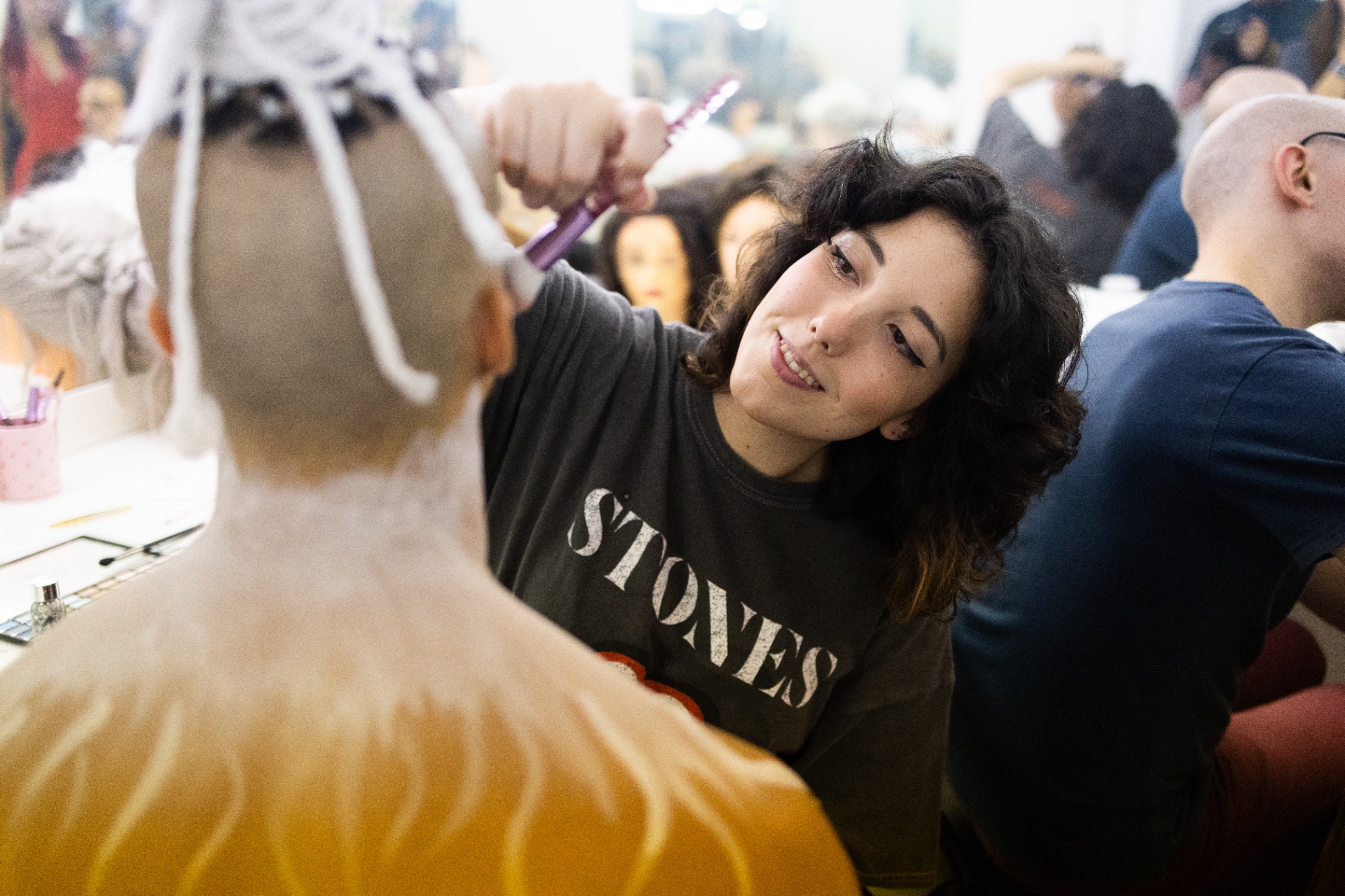 Truccatrice con t-shirt Stones sistema una parrucca bianca a un attore di spalle in camerino affollato