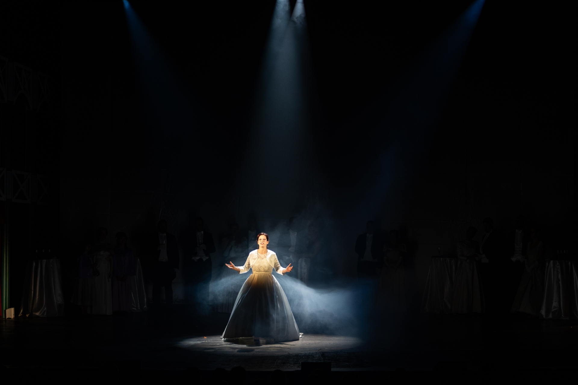 Cantante in abito bianco al centro del palco illuminata da tre fasci di luce, ensemble in ombra