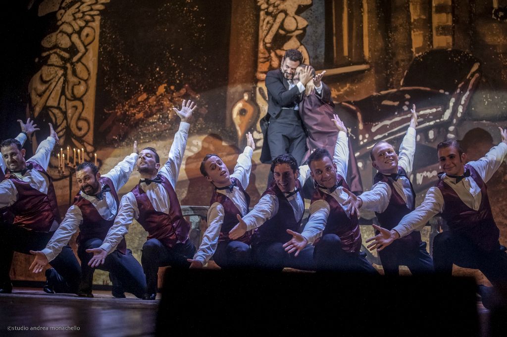 Coreografia di nove uomini in gilet bordeaux con braccia tese, due in piedi sullo sfondo