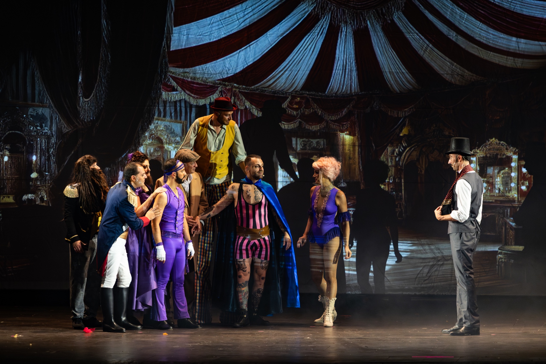 Il cast dei freaks radunato al centro sotto un tendone da circo dipinto, showman a destra