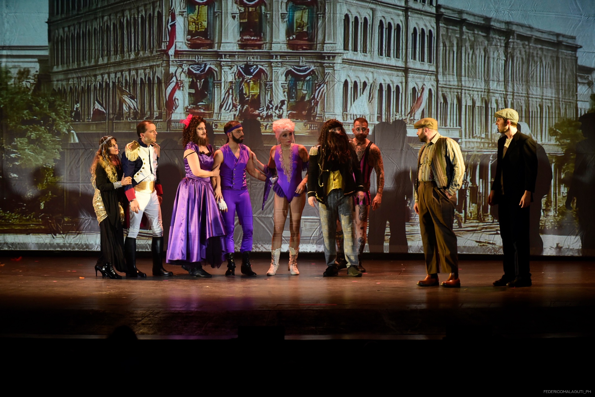 Cast in costumi viola davanti a una scenografia dipinta di edifici storici